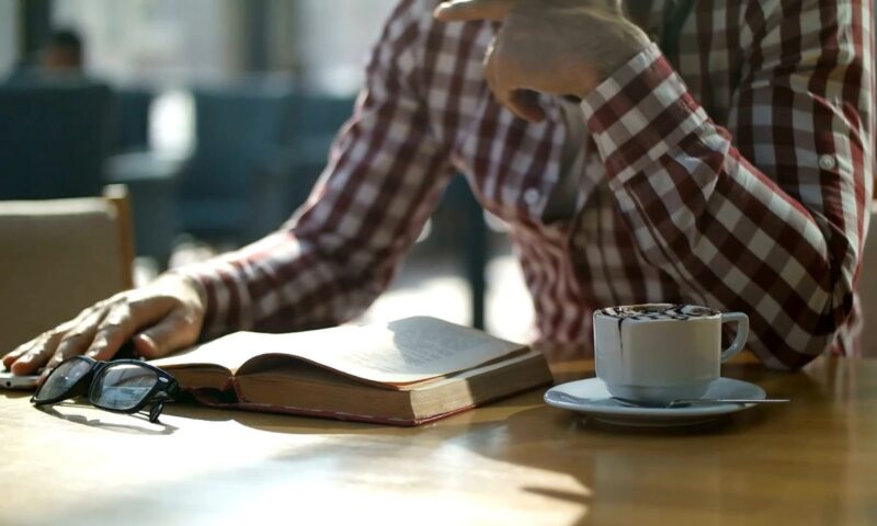 muška osoba sedi u kafiću, pije kafu i čita knjigu