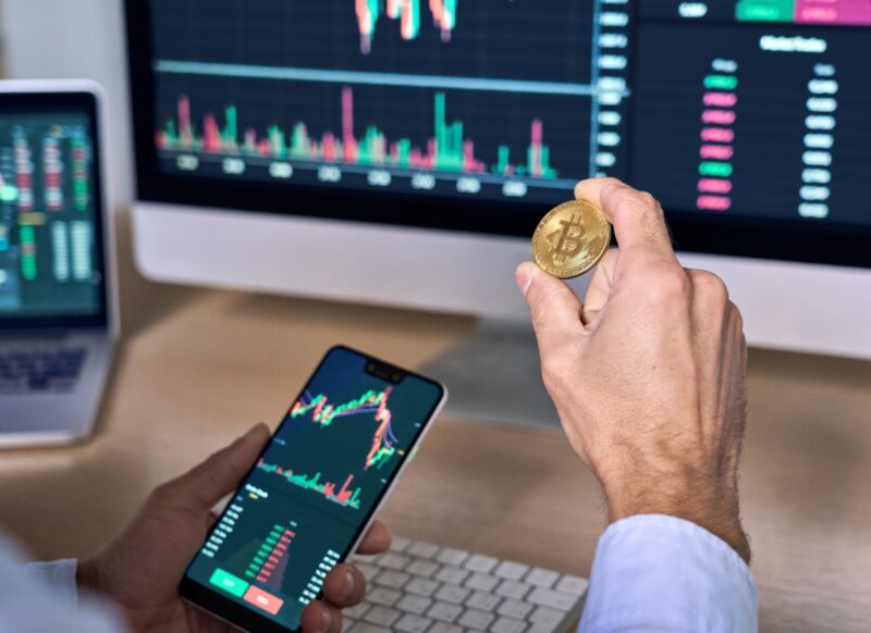 osoba proverava kriptovalute na telefonu i komjuteru i drži kripto novčić u ruci